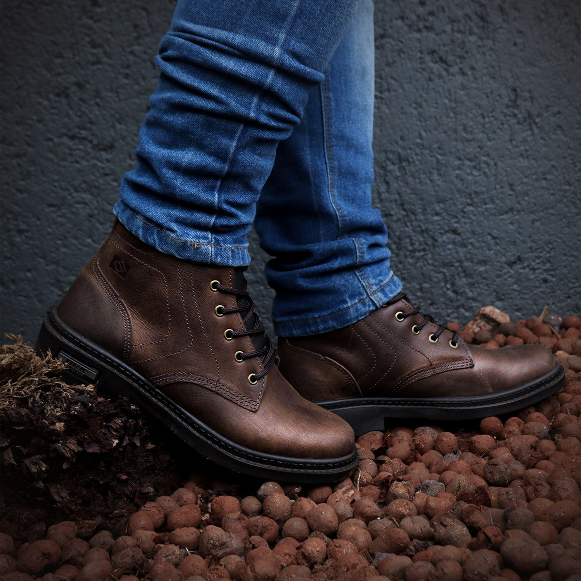Botas masculinas marrons com cadarço usadas com calça jeans azul, em chão com pedras e fundo cinza