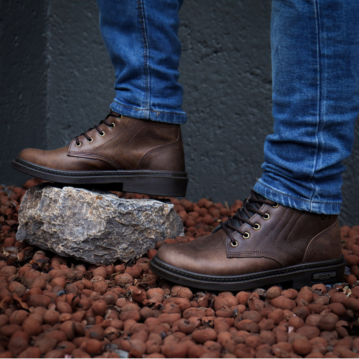 Botas marrons masculinas com cadarço usadas com jeans azul sobre pedras vermelhas e parede cinza