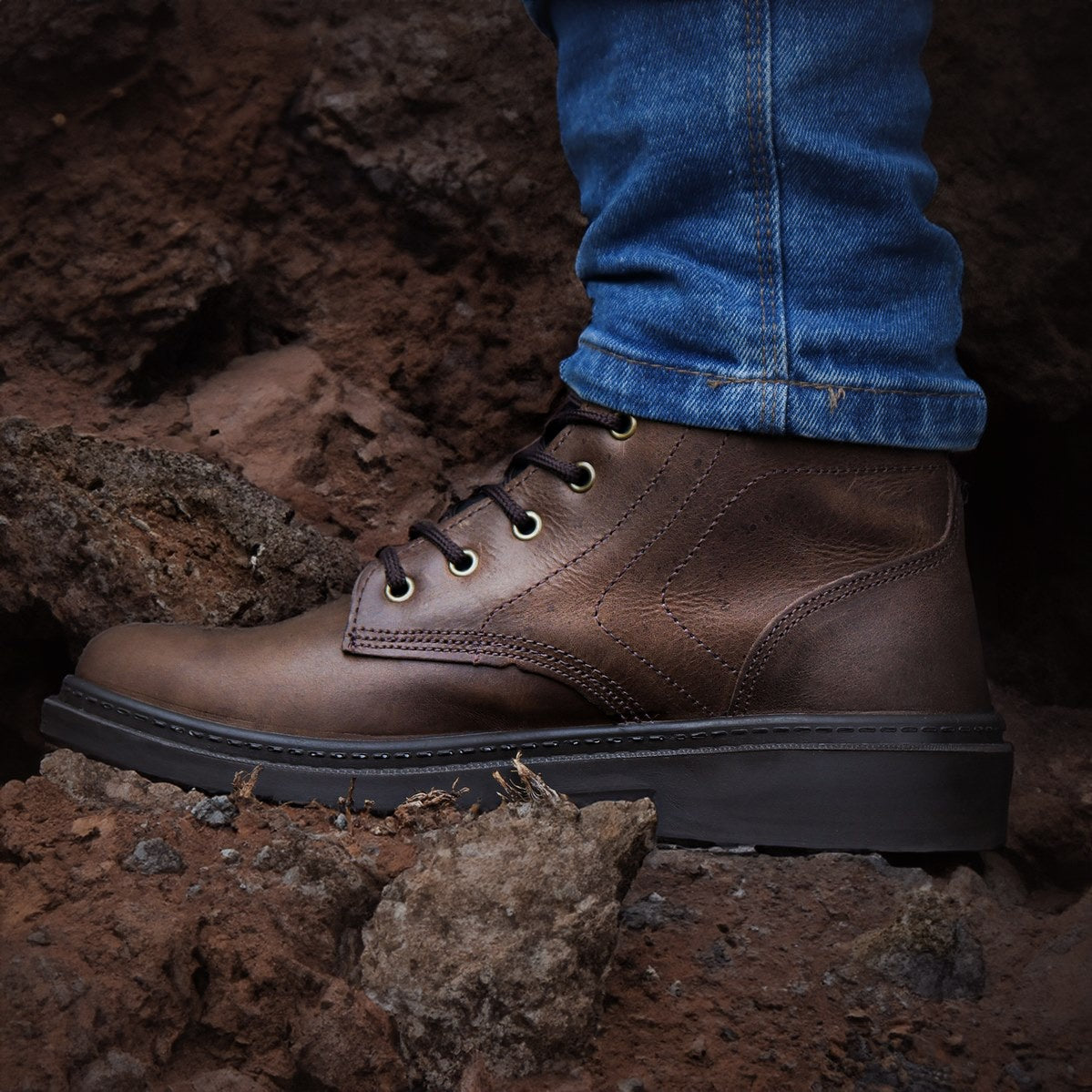Bota masculina marrom de couro com cadarço e calça jeans azul em terreno de pedras